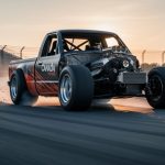 Low-angle view of a modified racing pickup truck speeding through a road-course corner with visible roll cage and tire traction.