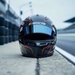 “A driver helmet placed at the edge of a NASCAR pit lane, with the racetrack pit area blurred in the background.”