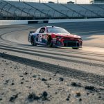 Low-angle view of a NASCAR stock car speeding on Nashville Superspeedway’s concrete banking with track texture and blurred grandstands in the background.