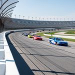 Stock cars speed through Talladega Superspeedway’s high-banked tri-oval in a tight drafting pack under sunny skies.