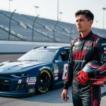Young NASCAR driver in a racing suit posing beside a Trackhouse-style stock car on a racetrack with blurred stadium and pit background.