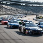 NASCAR stock cars run in a tight pack on Talladega Superspeedway’s steeply banked tri-oval during a full-throttle race under bright daylight.