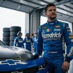 Alex Bowman in a Hendrick Motorsports firesuit stands beside a NASCAR stock car in the garage with blurred pit crew activity in the background.