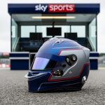 Close-up of a Formula 1 driver helmet placed on the pit lane with a blurred racetrack and broadcast booth background behind it.