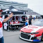 Canadian fan near a NASCAR stock car and merch tent at a track fan zone, with grandstands and crowd silhouettes in the background.