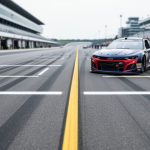 View of a NASCAR starting grid and pit lane at a race track, with an empty front-row area and a stock car in the foreground under an overcast sky.